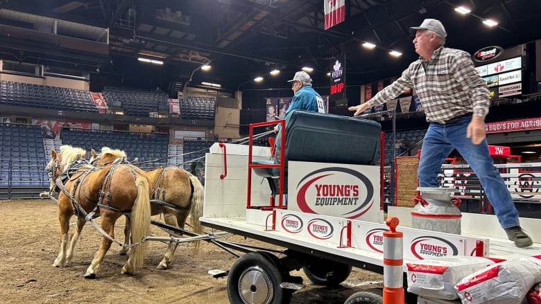 Man rides carriage guiding two horses in indoor riding competition, with a man behind him reaching out for his seat.