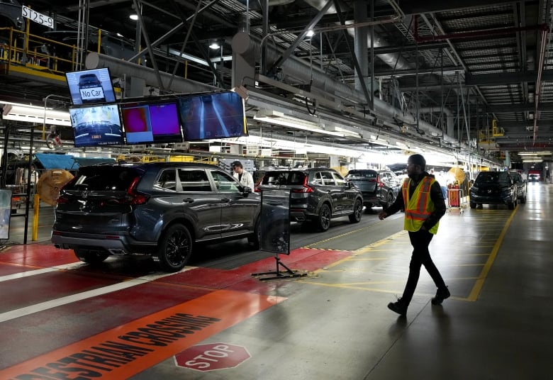 A man in a neon safety vest walks toward an auto assembly line where a worker in a white coat inspects SUVs on the line. 