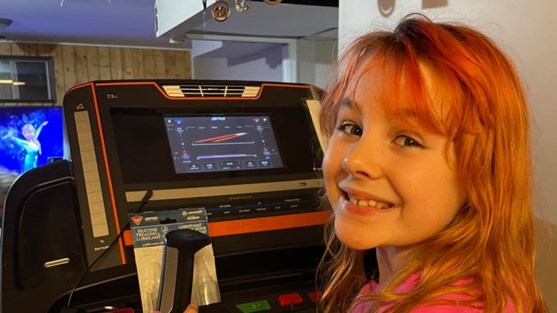 A young girl with dyed orange hair and wearing a pink sweater is on a treadmill and turning to the camera to smile for a picture. 