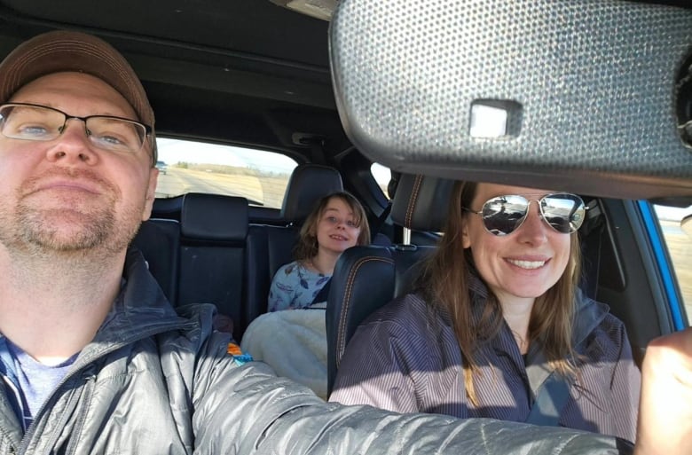 A family of three a mom and dad are in the front seat with their young daughter in the back seat posing for a picture smiling. 