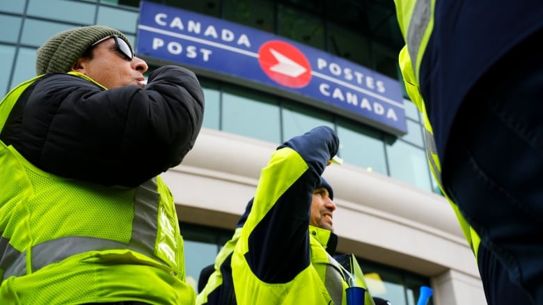 People in jackets and yellow vests stand outside a building with a sign reading 