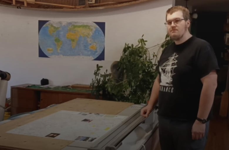 A man stands in front of a table with a map on it. A map of the world hangs on a wall behind him.