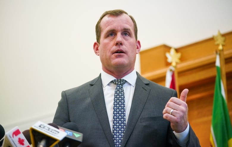 A man gestures with his hand. The man is wearing a grey suit, white shirt and blue tie. 