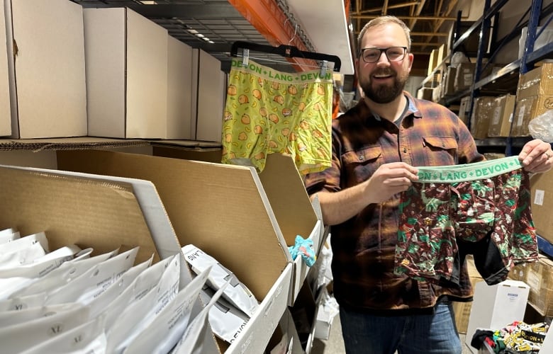 Tyler McCombs stands holding a pair of mens boxers next to a shelf of packaged orders ready to be shipped out.