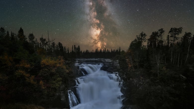 A photo taken at night of a bright milky way galaxy above a river and small waterfall in a forest.