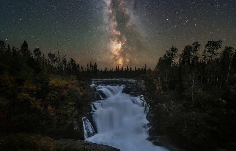 A photo taken at night of a bright milky way galaxy above a river and small waterfall in a forest.